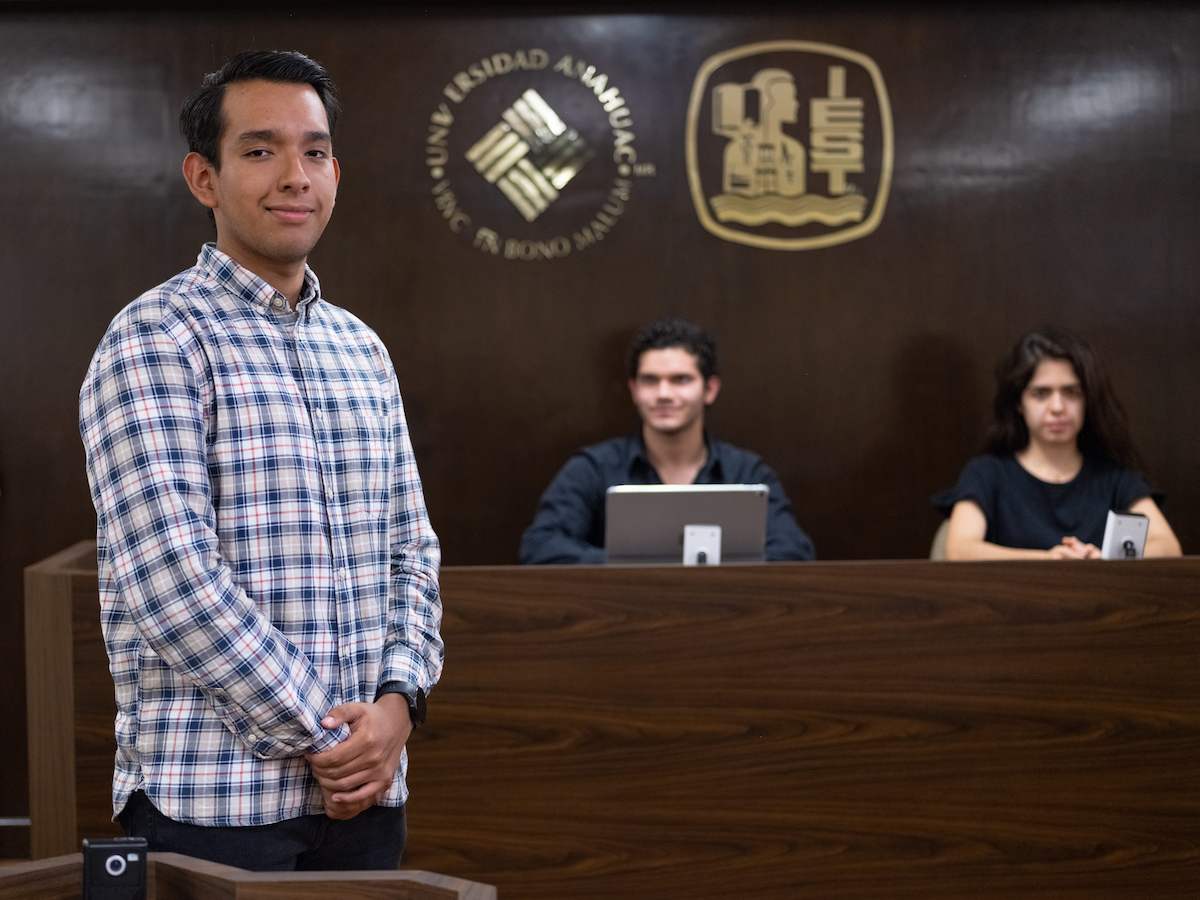 Estudiante de administración pública y gobierno en un aula tipo tribunal, acompañado de dos compañeros con laptops, reflejando formación profesional, liderazgo y toma de decisiones en contextos institucionales.