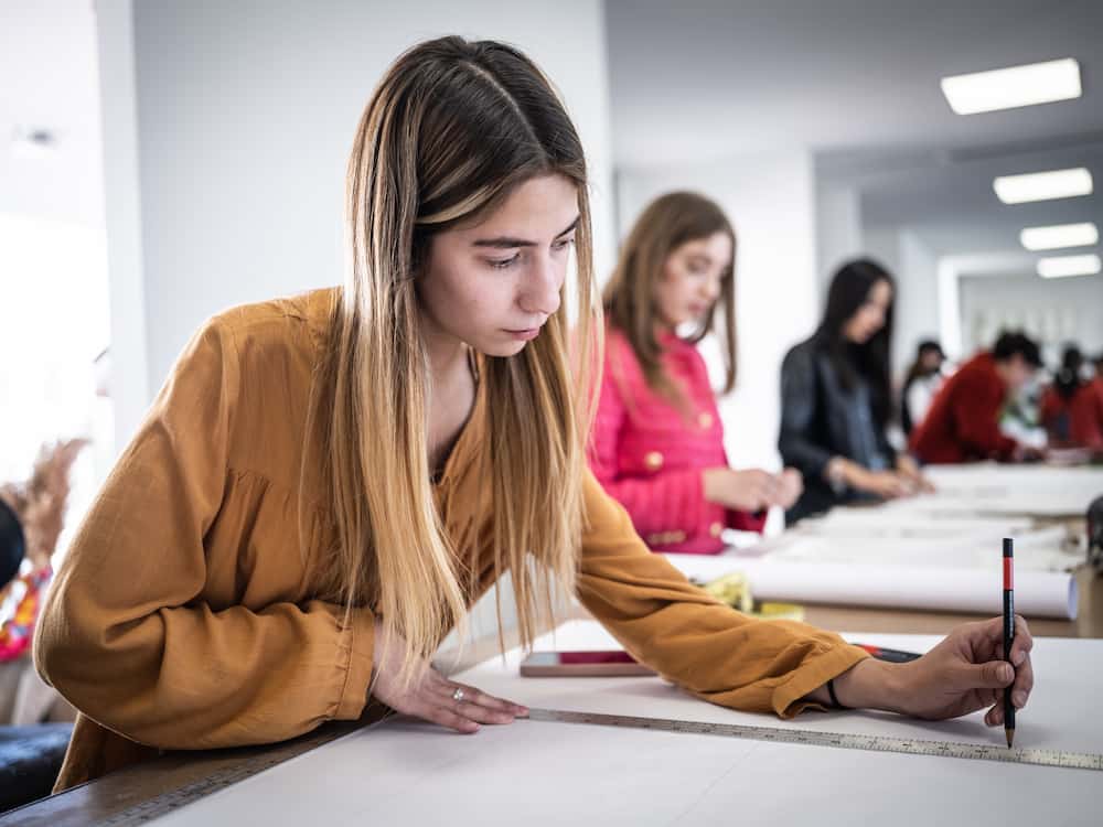 Estudiante trazando líneas con regla sobre papel en aula de diseño, con compañeras trabajando al fondo.