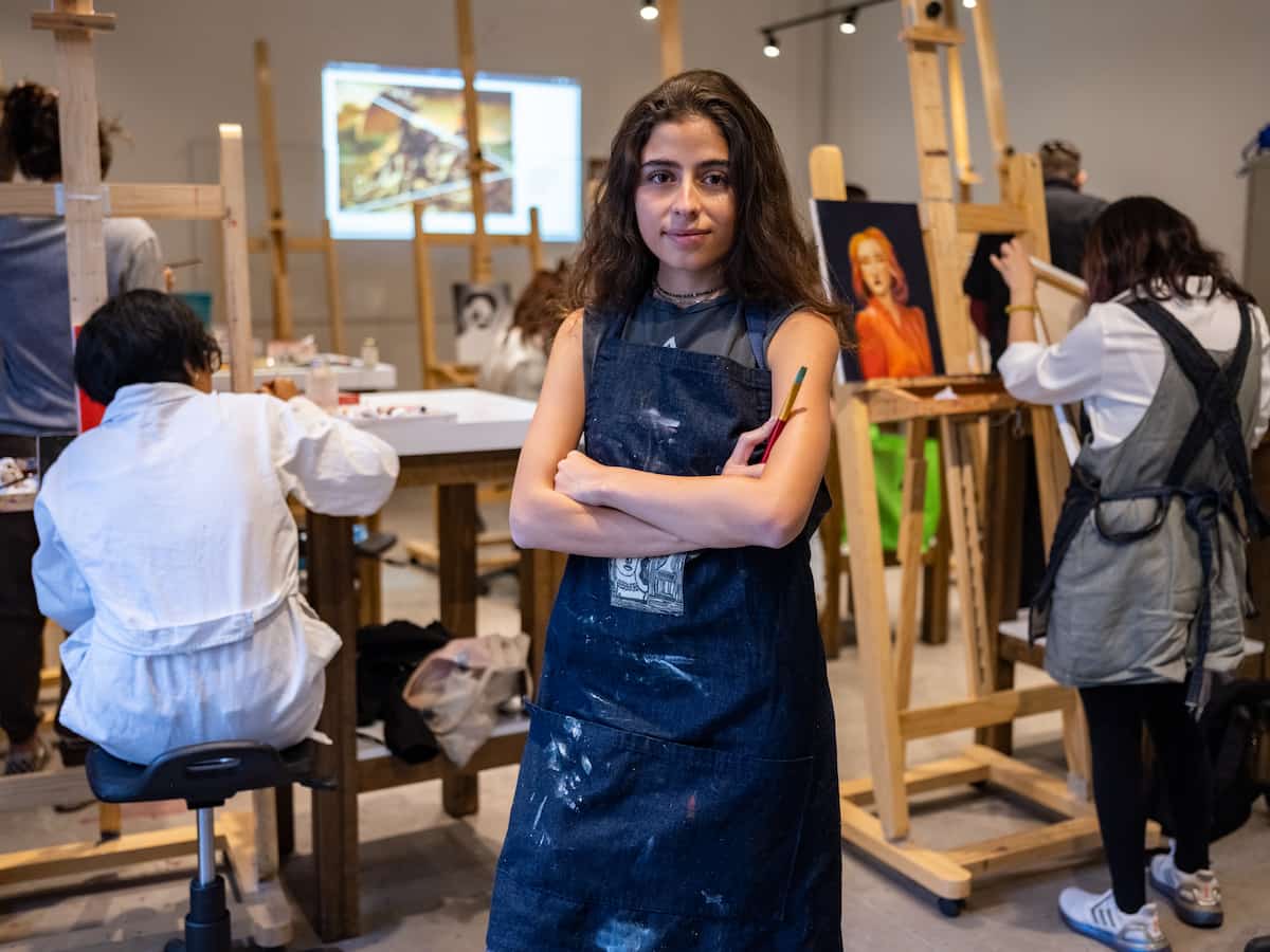 Joven artista en formación cruzada de brazos en aula de pintura, rodeada de estudiantes trabajando en sus obras.