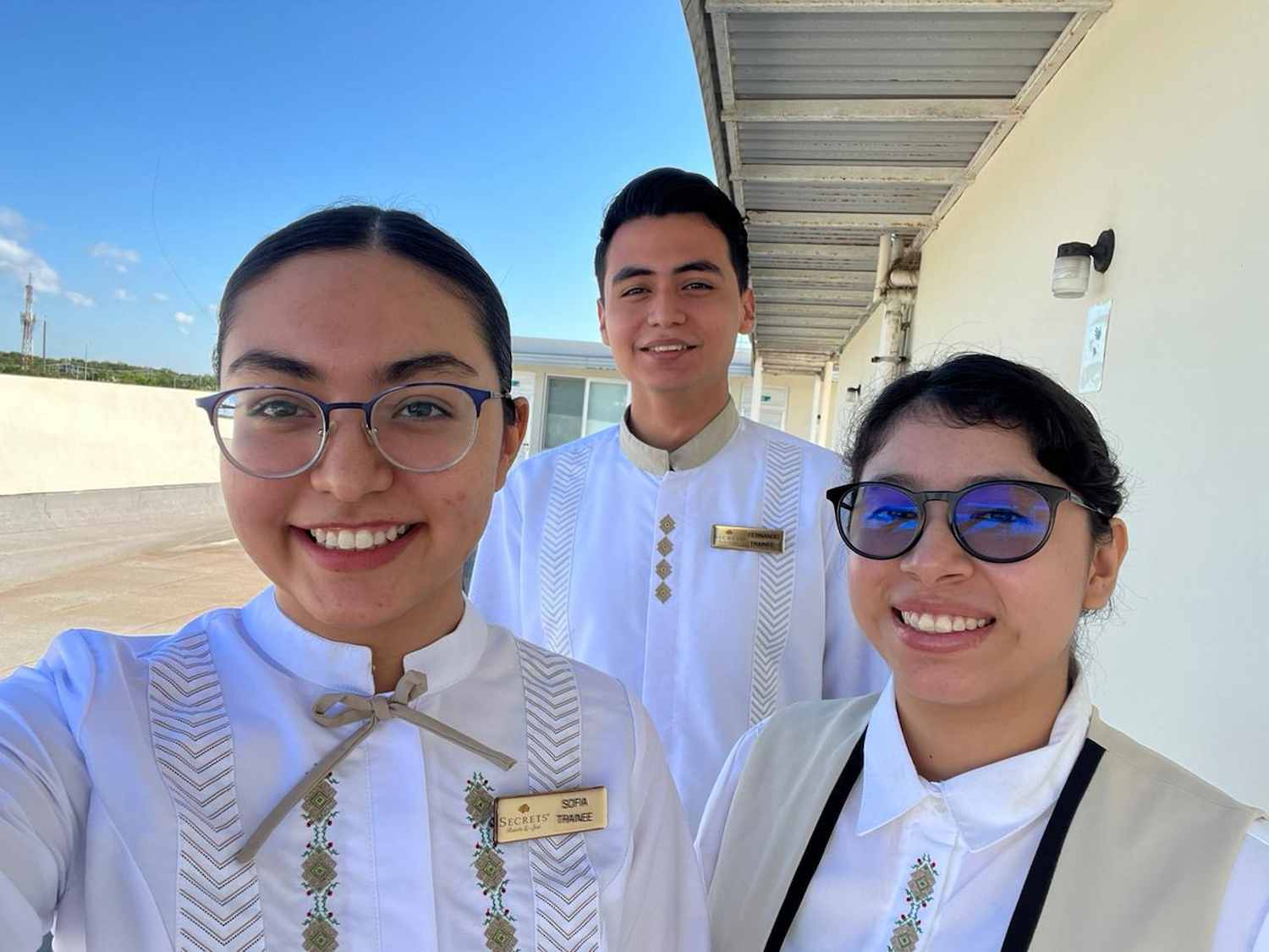 Tres jóvenes con gafete y uniforme blanco tradicional posan sonrientes al aire libre, bajo un techo metálico junto a una pared clara.