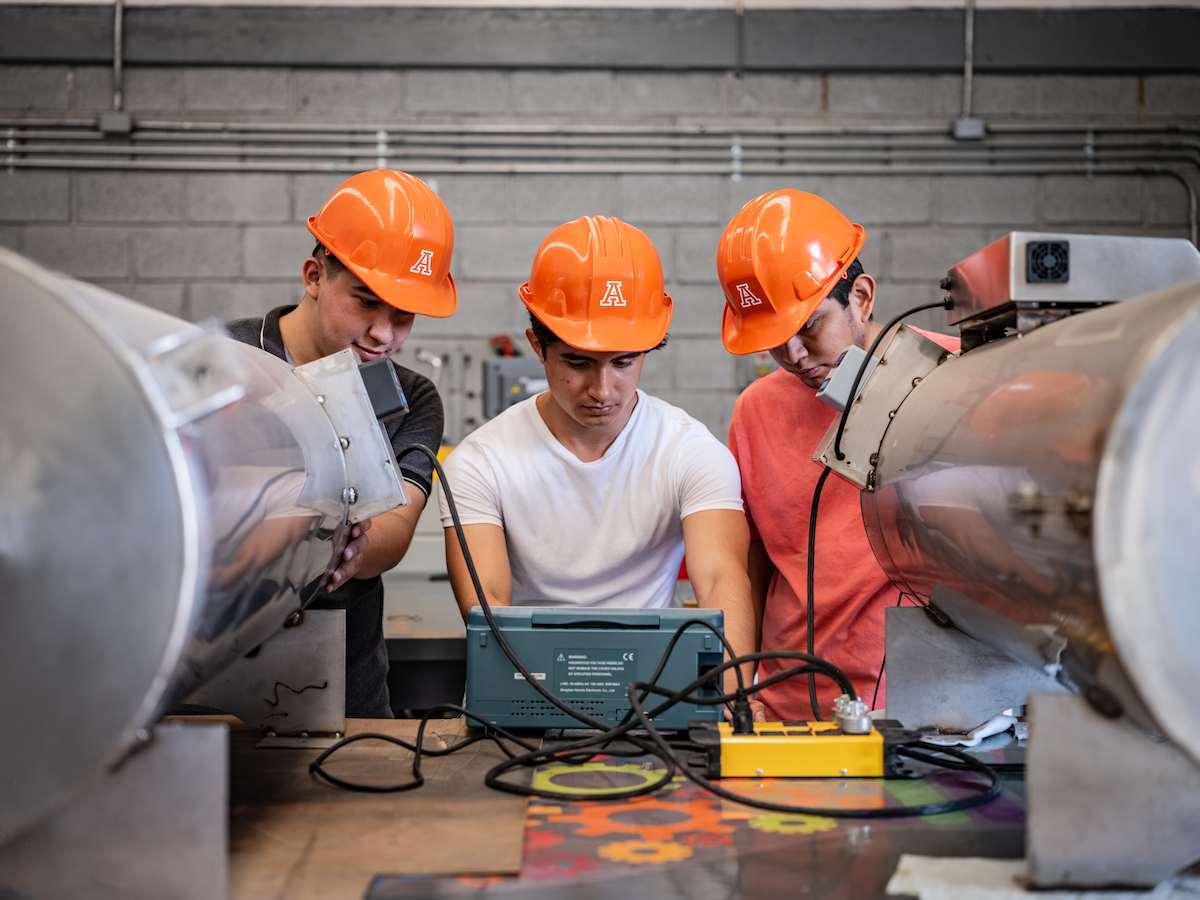 Tres estudiantes con casco naranja realizan pruebas con equipo electrónico conectado a grandes tubos metálicos, dentro de un laboratorio industrial.