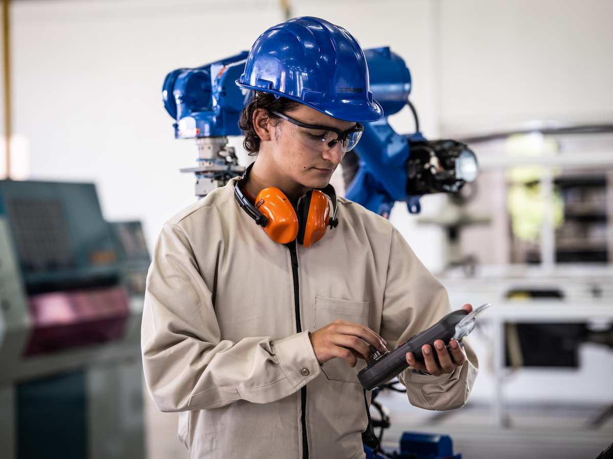 Estudiante con casco azul y protección auditiva opera un control de mando frente a brazo robótico industrial en laboratorio de automatización.