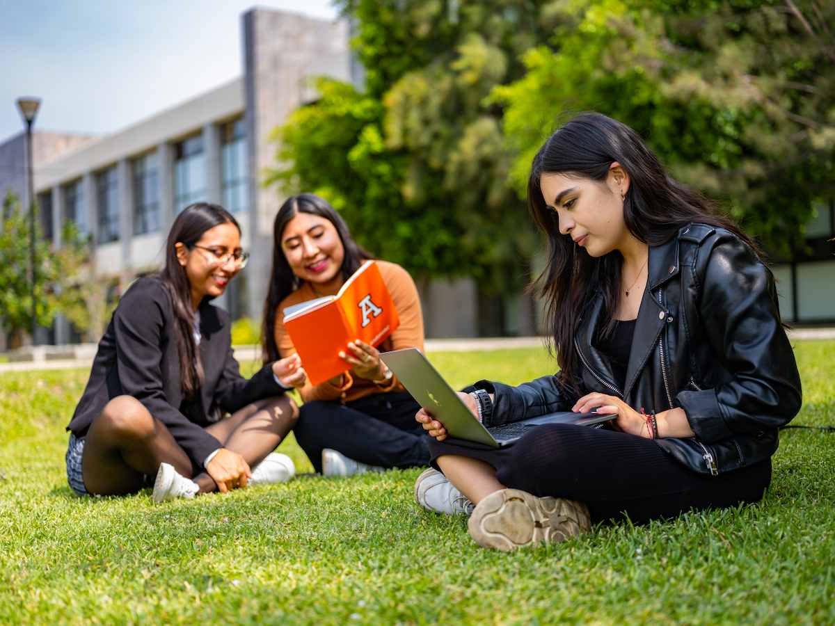 Tres estudiantes de Lenguas y Gestión Cultural estudiando juntas al aire libre, usando una laptop y un cuaderno, reflejando trabajo colaborativo, aprendizaje de idiomas y enfoque en la diversidad cultural.