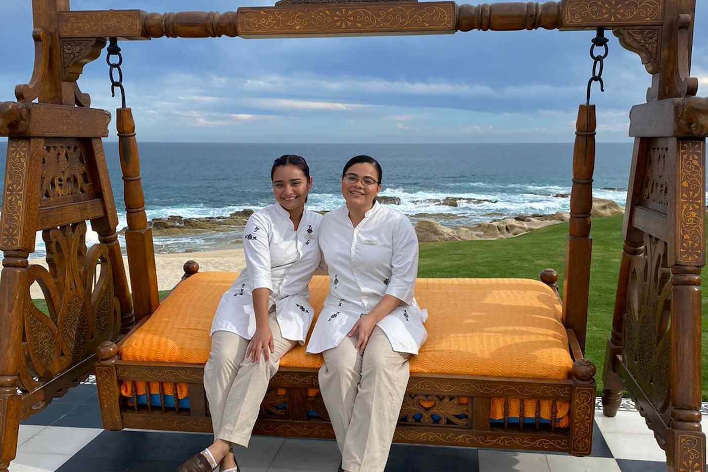 Dos chefs sentadas relajadamente en un columpio tallado con vista al océano en un entorno costero.